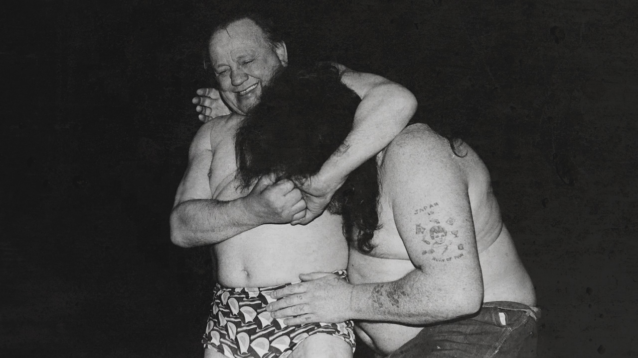 Les Kellett has another wrestler in a headlock, with his long hair covering his face and Kellett smiles