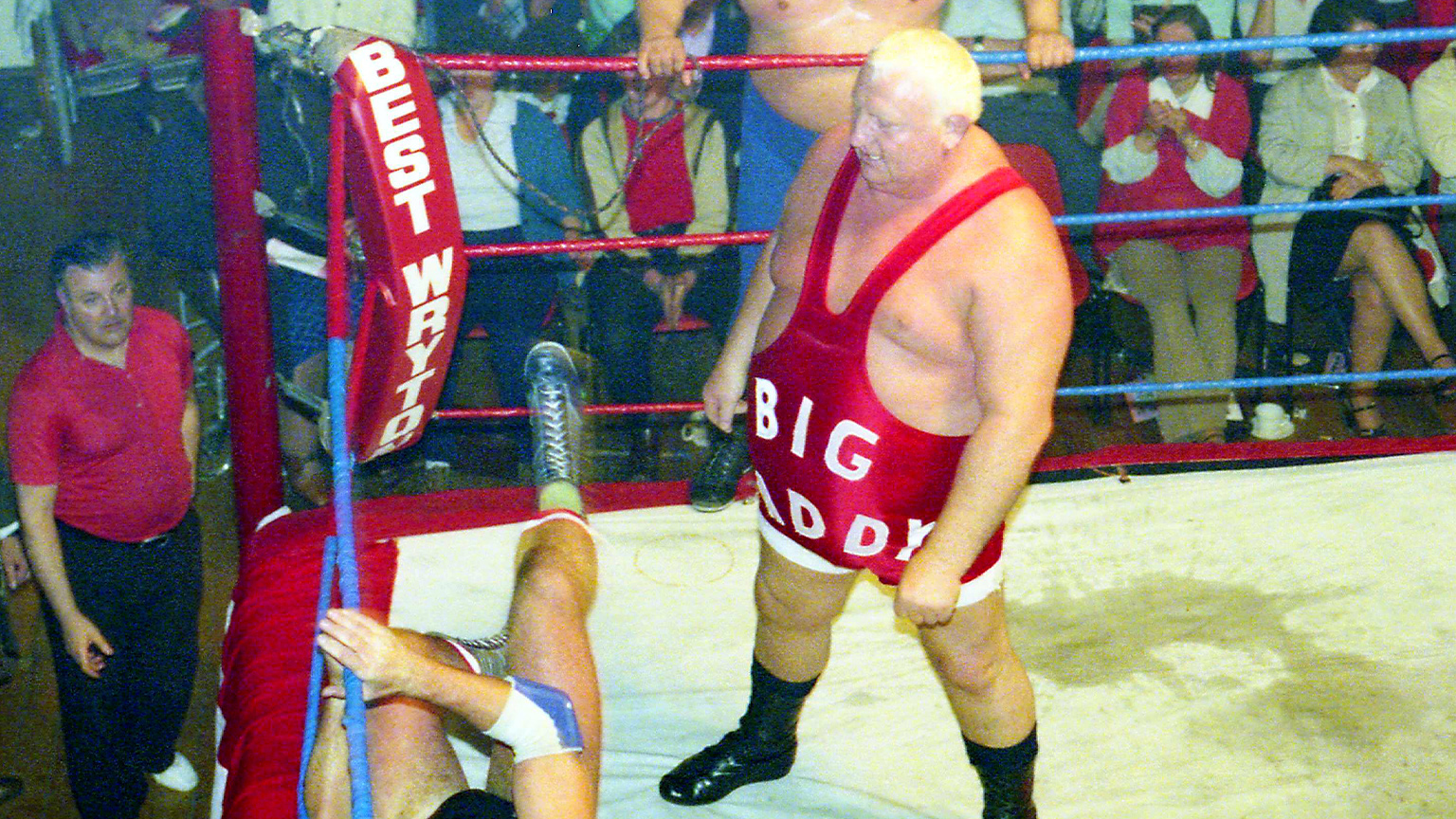 Big Daddy in a red singlet with "Big Daddy" in white letters wrestling in a match for Best Wryton promotions. A wrestler is on the floor while King Kong Kirk is standing on the apron.