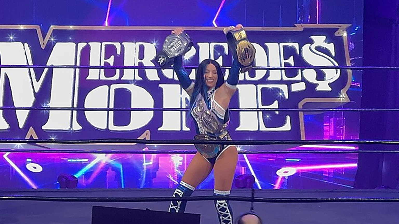Mercedes Mone standing in the ring holding up numerous title belts as her name is on the screen behind her.