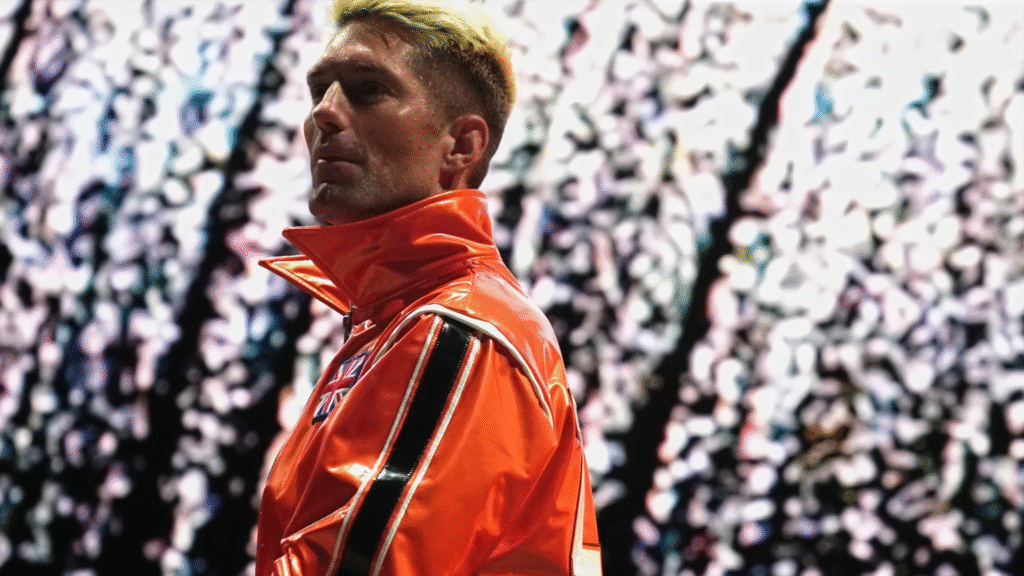 A close up of Zack Sabre Jr wearing orange as white fireworks go off behind him.