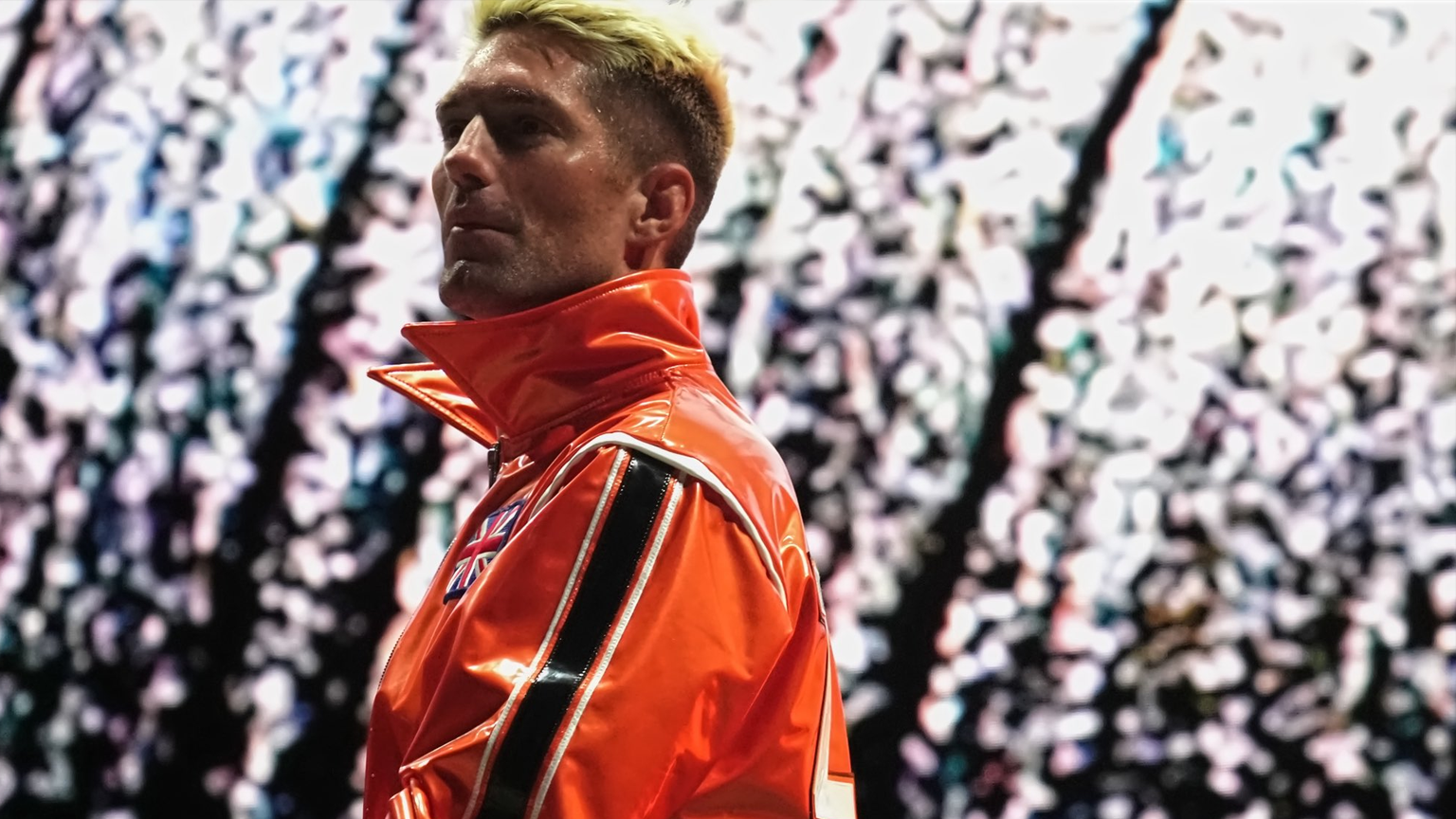 A close up of Zack Sabre Jr wearing orange as white fireworks go off behind him.